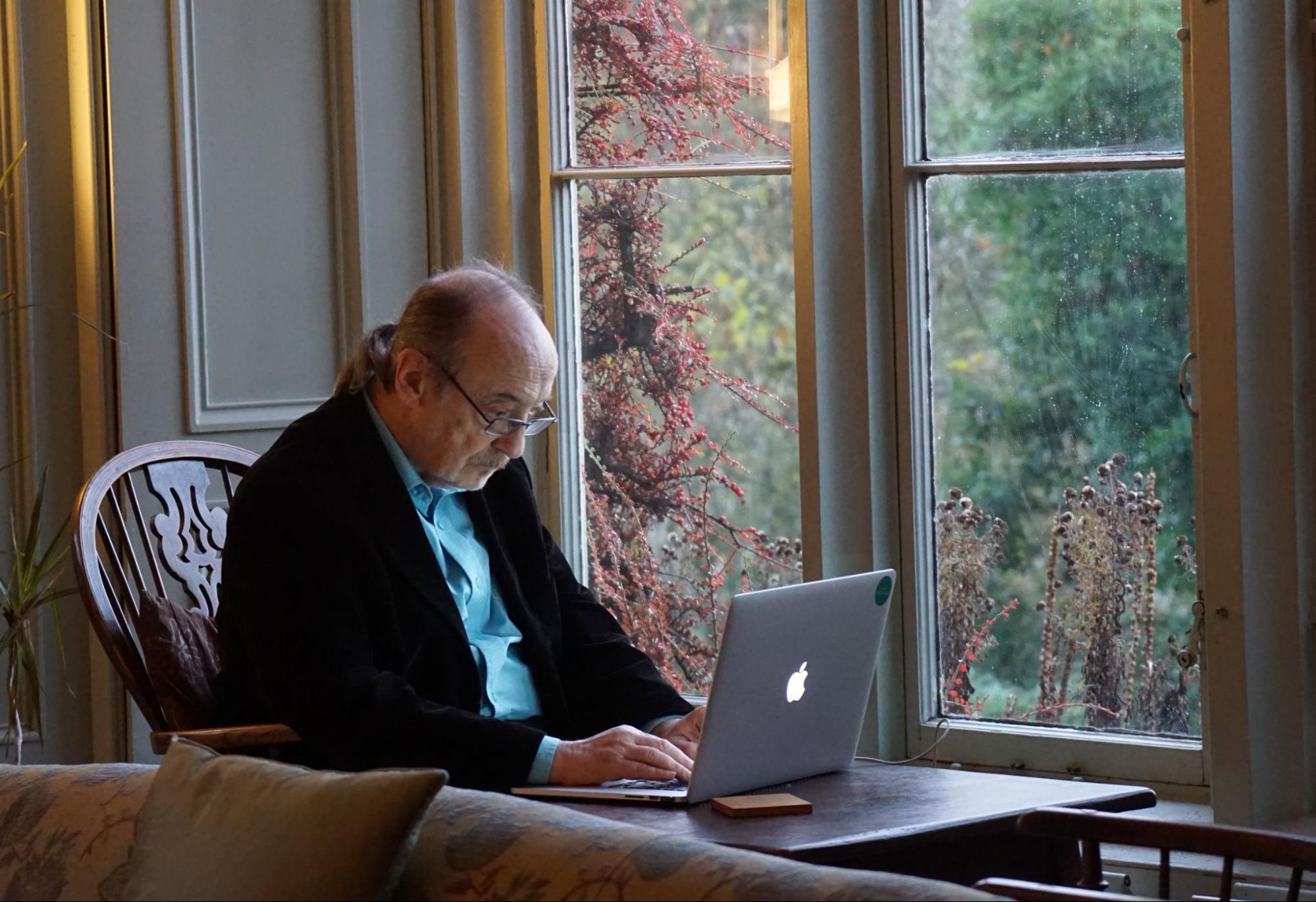 An older man sitting by the window at a laptop.