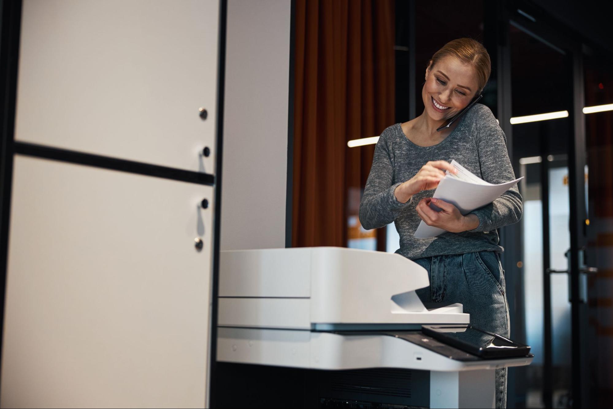 A woman on a phone smiling in front of a printer with paper sheets.